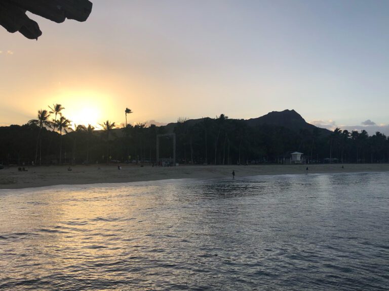 A photo of Diamond Head, taken at sunrise. The mountain is in a silhouette and the sun is on the left side. There is ocean in the foreground. The beach is Waikiki Beach.